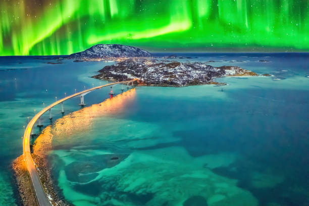 Stunning aerial view of the Sommaroy Bridge in Tromso, Norway, illuminated at night under the glowing Northern Lights. The vibrant aurora dances across the sky, reflecting on the calm turquoise waters below, with snow-covered hills in the background. This breathtaking natural phenomenon is set against a remote arctic seascape.